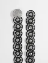 Cinturón ancho de cuero sintético negro, compuesto por secciones circulares unidas, decorado con múltiples ojales metálicos plateados y un dije central grande con relieve de un jinete a caballo. El cierre es mediante un broche metálico grande con el mismo diseño de relieve.