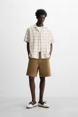 Camisa de manga corta color crudo con estampado de cuadros en tonos grises y marrones. Tiene cuello bowling, corte relaxed fit, aberturas laterales en el bajo y cierre frontal con botones.