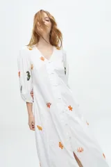 Vestido midi color arena con escote en V, mangas 3/4 abullonadas y cierre frontal con botones. Presenta bordados de motivos marinos.