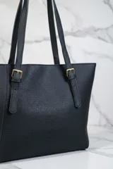 Bolso tipo tote negro de material texturizado, con dos asas de hombro ajustables mediante hebillas doradas.