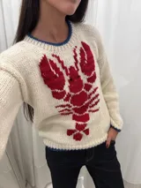 Sweater tejido a mano en Uruguay, confeccionado en lana merino color crudo, con diseño de langosta roja en el frente y detalles en azul en cuello y puños.