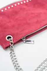 Cartera de mano o clutch color rojo oscuro (bordó) con textura de gamuza o terciopelo. Presenta tachas metálicas plateadas a lo largo del borde inferior y lateral, y un cierre perimetral plateado. Incluye una correa de hombro desmontable compuesta por una tira de material a tono y cadena metálica plateada.