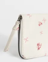 Billetera de cuerina color crema con estampado de flores y moños. Tiene formato rectangular, cierre circundante con tirador y múltiples compartimentos internos.