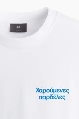 Remera blanca de algodón de peso medio con cuello redondo ribeteado, hombros caídos y bajo recto. Tiene un estampado pequeño en el lado izquierdo del pecho con texto en griego de color celeste. Corte holgado.