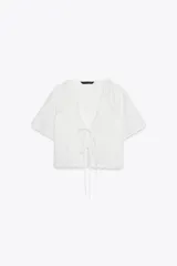 Camisa blanca de manga corta con escote en pico y cierre frontal con lazadas. Presenta bordados florales a tono.