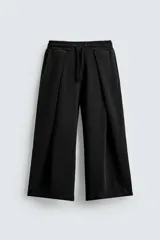 Pantalón jogger negro de corte relaxed fit, confeccionado en tejido de algodón. Presenta cintura elástica ajustable con cordón, pliegues frontales, bolsillos delanteros y un bolsillo trasero.