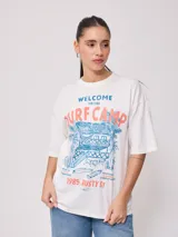 Remera blanca de algodón con estampado frontal de estilo playero con texto "Welcome to the Surf Camp" y dibujo de una casa de playa con palmeras y tablas de surf.