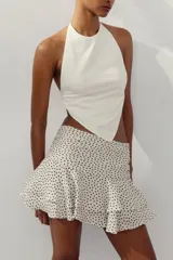 Falda pantalón corta color crudo con estampado de lunares negros, tiro medio, detalle de pliegues y volantes, y cierre lateral con cremallera.