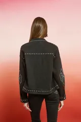 Campera de jean negra de corte holgado con detalles de tachas plateadas en costuras, hombros y puños, y bordado floral blanco en las mangas.