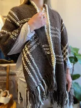 Ruana tejida en lana color marrón con detalles geométricos en blanco y flecos en el ruedo.