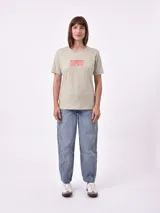 Remera blanca de algodón con cuello redondo y manga corta. Presenta un pequeño bordado en el pecho izquierdo con una figura que parece ser una persona acostada en una tabla de surf o flotador.