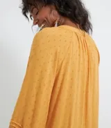 Blusa de manga larga color mostaza con textura de puntos en relieve (dobby). Presenta cuello redondo con abertura en V y cordón ajustable con borla (tassel) en el escote. Las mangas tienen detalles de entretejido calado (entremedios) y volados con terminación fruncida en los puños.