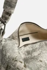 Bolso de hombro color gris con efecto desgastado, fabricado en tejido de algodón. Tiene un compartimento principal con cierre de cremallera, un bolsillo interior con cremallera y asa de hombro ajustable.