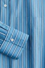 Camisa celeste y blanca a rayas verticales, de corte amplio y mangas largas. Tiene cuello clásico, cierre frontal con botones y bolsillo de parche en el pecho.