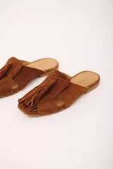 Zueco de cuero gamuzado color marrón, con punta cuadrada y detalle de gran borla de flecos en el empeine. Presenta aberturas laterales y suela plana.