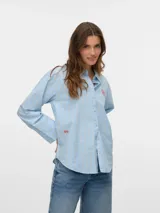 Camisa celeste a rayas verticales blancas, de corte amplio, manga larga con puños abotonados, cuello clásico y cierre frontal con botones. Presenta un bordado de una flor roja en el pecho y dos corazones rojos en el puño.