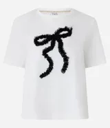 Remera blanca de manga corta con cuello redondo y un aplique frontal de lazo tridimensional hecho con tul negro fruncido.