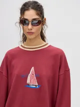 Buzo blanco de corte redondo (crew neck) con puños ajustados que tienen rayas rojas y blancas. Presenta un estampado central de un velero navegando con texto alrededor.