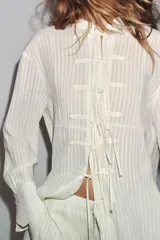 Camisa blanca a rayas verticales, de cuello redondo y manga larga con puños. Presenta cierre frontal con cordones ajustables con lazadas y abertura en la espalda ajustable con lazadas.