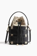 Cartera bombonera de cuero sintético color beige con apliques de tiras de cuero sintético negro y costuras blancas. Tiene doble asa corta y correa larga extraíble. El interior es de lona con cordón ajustable.