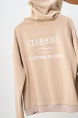 Conjunto deportivo color beige compuesto por un canguro con capucha y un pantalón jogging. El canguro tiene un bolsillo tipo canguro y estampados en el frente. El pantalón es de corte recto con cintura ajustable con cordón.