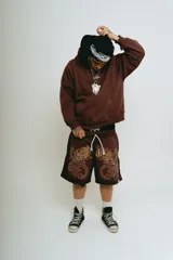 Bermuda de felpa color marrón oscuro con efecto lavado (washed) y detalles en negro en los laterales. Presenta un gran estampado serigrafiado en dorado con tipografía oriental y motivos ornamentales. Tiene cordón de ajuste en la cintura y remaches metálicos cromados en los laterales.