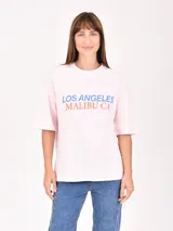 Remera rosa de corte holgado con estampado frontal que dice 'Los Angeles Malibu CA' en azul y naranja.