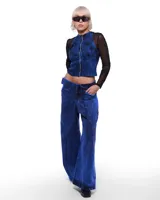 Pantalón de denim azul intenso, corte sastrero de pierna ancha, tiro alto o a la cadera (según el talle elegido), cierre frontal invisible, pretina cruzada y bordado negro del logo Temblad en la parte trasera.