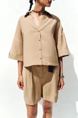 Camisa color beige de cuello solapa con escote pico y manga corta ancha acabada en vuelta con trabilla y botón. Cierre frontal con botones.