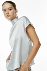 Blusa color crudo de tejido satinado, con cuello subido, manga sisa y cierre en la espalda con lazada y botones.