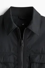 Campera negra con cuello camisero, cierre frontal y bolsillos superiores con solapa. Elástico revestido en puños y bajo.