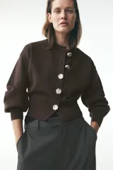 Chaqueta de punto marrón oscuro con cuello redondo y manga larga abullonada. Cierre frontal con botones metálicos plateados con diseño de rosa.