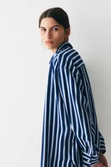 Camisa oversized de sarga a rayas verticales azules y blancas, con cuello de pico y botonadura frontal. Modelo con hombros caídos, mangas largas con tapeta y puños abotonados, y canesú en la espalda. Bajo recto con aberturas laterales redondeadas.
