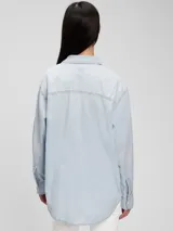 Camisa oversize de jean celeste claro, con cuello clásico, cierre frontal con botones a presión, dos bolsillos con solapa en el pecho y mangas largas con puños abotonados.