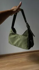 Cartera negra de lona semi impermeable con cierre metálico, forrería de gross y bolsillo interno. Tiene tira regulable y etiqueta blanca con texto en el frente.