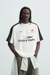 Camiseta blanca de corte holgado con mangas cortas negras. Presenta un logo azul en el pecho izquierdo, un escudo rojo con un caballo en el pecho derecho y la inscripción "Victory Games" en negro en el centro.