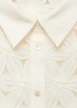 Blusa color crema con textura calada y flores en relieve, cuello camisero, manga corta y cierre de botones en el cuello.