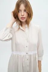 Vestido camisero midi color beige a rayas verticales, con cuello mao, mangas 3/4 abullonadas y festones de algodón en el cuerpo. Cierre frontal con botones.
