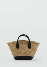 Bolso tipo canasta de paja natural con base y asas cortas de cuero sintético negro.