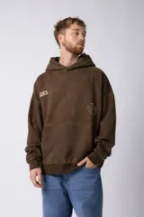 Buzo canguro marrón de frisa rústica con capucha y corte oversize. Presenta un bolsillo tipo canguro y estampados con el logo de la marca en el frente y la manga.