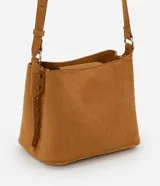 Cartera de gamuza color tostado, de tamaño mediano, con diseño transversal y asa larga ajustable. Cuenta con cierre superior de botón a presión, compartimento interno principal y detalles laterales con ojales metálicos.