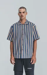Camisa de manga corta con cuello abierto y calce oversize confeccionada en textil sarga liviana con estampado a rayas verticales en azul, naranja y amarillo.