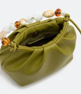 Bolso pequeño color verde oliva, desestructurado, con pinzas frontales y cierre de botón a presión. Incluye una correa de hombro extraíble y un asa de mano decorada con cuentas acrílicas variadas en tonos blancos, marrones y transparentes.