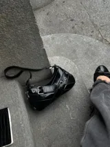 Cartera tipo baguette negra de cuero sintético con correa corta y detalles de hebillas plateadas.