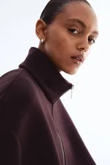 Campera corta de tejido efecto neopreno color marrón oscuro o ciruela, con cuello chimenea alto y cierre frontal metálico. Presenta hombros caídos y un bajo fruncido con elástico que le da un calce holgado.