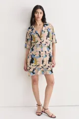Vestido camisero corto con estampado abstracto en tonos beige, azul y negro. Confeccionado en lino y seda, con cuello camisero, mangas cortas y cierre frontal con botones.