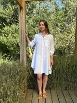 Vestido camisero blanco de lino, con cuello camisero, manga larga con botón para recoger, cierre frontal con botones de nácar, bolsillo en el pecho y diseño asimétrico más largo en la parte trasera.