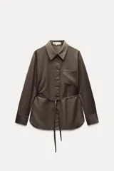 Camisa marrón de Zara Woman Collection, confeccionada en hilatura con viscosa. Tiene cuello solapa, manga larga con puño, bolsillo de plastrón en el pecho, cinturón ajustable con lazadas y bajo asimétrico. Se cierra con botones frontales.