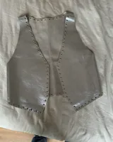 Chaleco corto plateado de cuero ovino con tachas metálicas en el cuello y el ruedo.