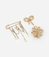 Kit de tres broches metálicos con baño dorado. Incluye un broche con forma de lazo texturizado, uno con forma de flor calada y un tercero tipo alfiler de gancho con cadenas colgantes que incorporan dijes de corazones y estrellas.